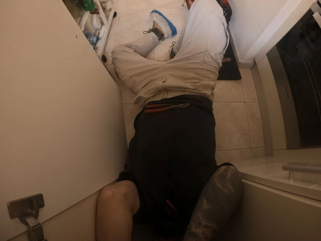On peut voir un plombier en dépannage allongé au sol qui travail sous un évier de cuisine